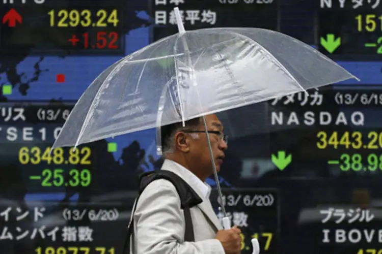 Homem segurando um guarda-chuva passa por um quadro eletrônico mostrando as cotações do índice Nikkei e de outras bolsas do mundo, em Tóquio no Japão (Issei Kato/Reuters)