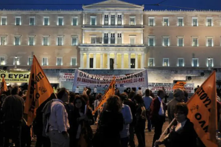 Manifestantes agitam bandeiras e exibem faixas durante protesto contra a aprovação da lei de reestruturação do setor público em frente ao Parlamento da Grécia, em Atenas (AFP / Louisa Gouliamaki)