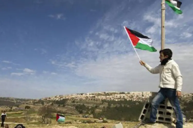 
	Jovem levanta uma bandeira palestina e pessoas rezam durante um protesto contra as col&ocirc;nias de Israel: o pa&iacute;s rejeitou de modo imediato as conclus&otilde;es (Ahmad Gharabli/AFP)