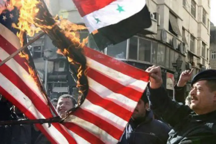 Com uma bandeira da Síria ao fundo, manifestantes queimam uma dos EUA em um recente protesto nas ruas de Damasco (Louai Beshara/AFP)