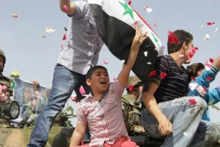 Sob uma chuva de pétalas de rosas lançadas por mulheres, jovens e crianças sobem em um tanque de guerra durante protesto na cidade de Daraa, na Síria (Louai Beshara/AFP)