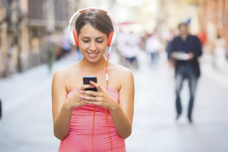 Mulher parada no meio da calçada usando smartphone e fones de ouvido (william87/Thinkstock)