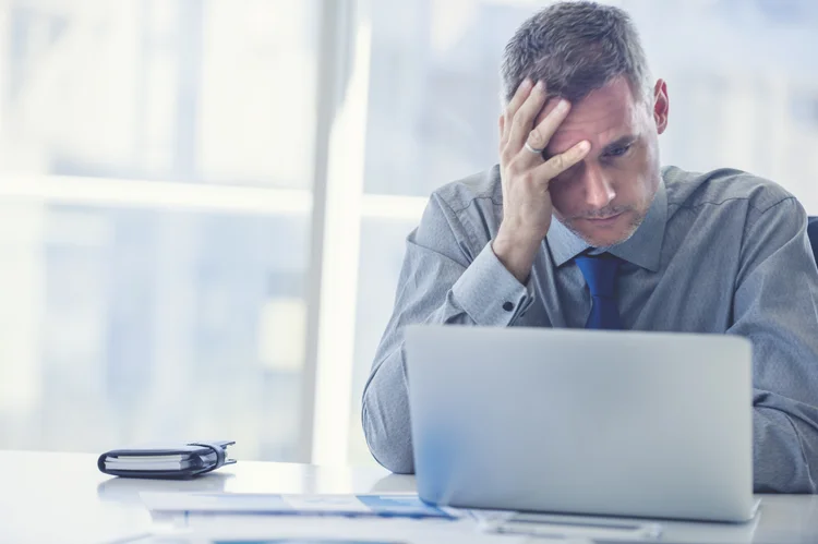 Stressed mature businessman with laptop. He could also have a headache. He is sitting in the boardroom. There are documents on the table. Copy space (courtneyk/Getty Images)