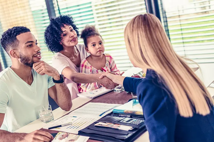 Female insurance agent in full suit sitting behind a desk  with african american family and handshaking with african american woman. (Agência/Getty Images)