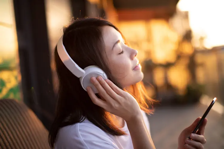 Portrait of smiling young Asian woman with eyes closed enjoying music over headphones and using smartphone, relaxing on the balcony against sunlight. (Getty Images/Getty Images)
