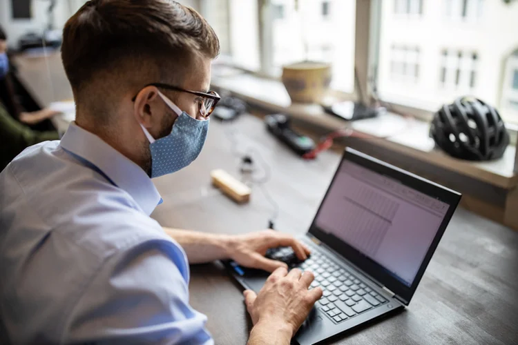 Close-up of a businessman with protective face mask working on his laptop at his desk. Male professionals return back to work after pandemic lockdown. (Luis Alvarez/Getty Images)