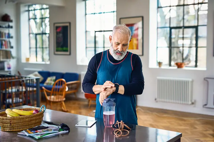 Active man in his 50s setting smart wear device, health drink on kitchen counter, checking data on digital running tracker, checking the time, getting ready for morning run (Reprodução/Getty Images)