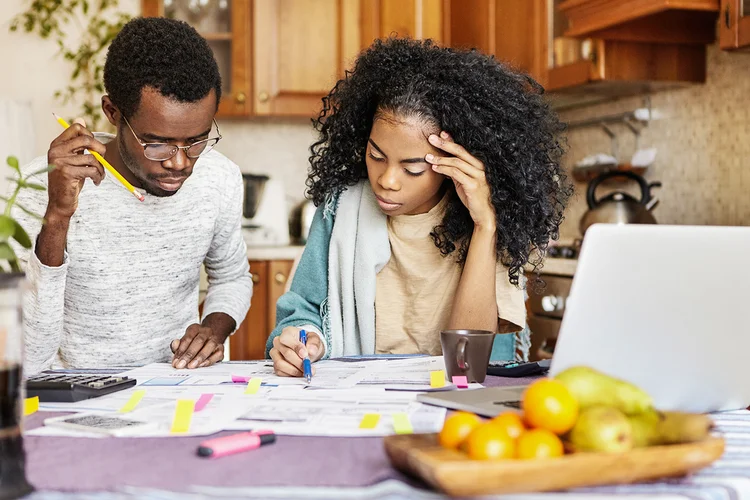 Young African husband and wife doing paperwork together at home, planning new purchase, calculating family expenses, sitting at kitchen table with laptop and calculator. Domestic budget and finances (Freepik/Divulgação)