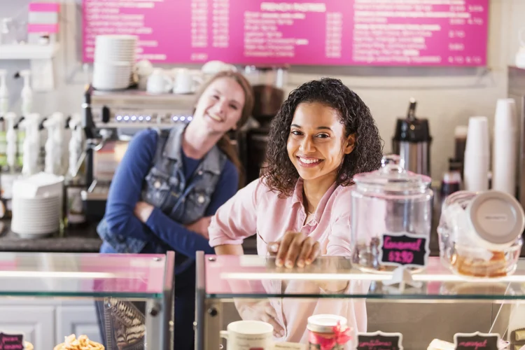 Two young multi-ethnic women working in a local coffee or pastry shop standing behind the display case, smiling at the camera. The focus is on the mixed race African-American and Caucasian woman in the foreground. (kali9/Getty Images)