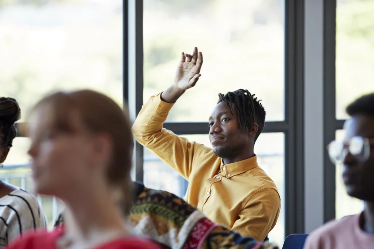 Confident young male student looking away while sitting with arm raised amidst friends in classroom against window at community college (Klaus Vedfelt/Getty Images)