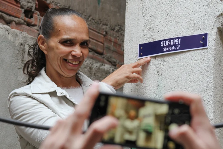 Alexandra Pereira Silva, de 46 anos, ao lado de placa com o seu primeiro endereço que aparece no Google Maps (André Silva/ Cria Brasil/Reprodução)