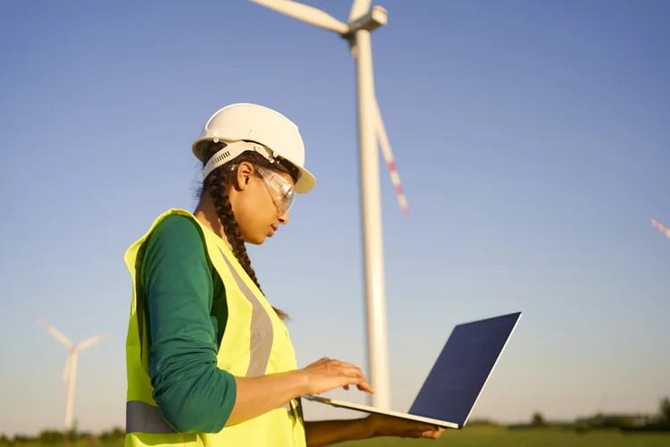 Female engineer wearing hard hat and reflective green jacket standing with laptop against wind turbine and setting it up. Energy concept. (Andriy Onufriyenko/Getty Images)