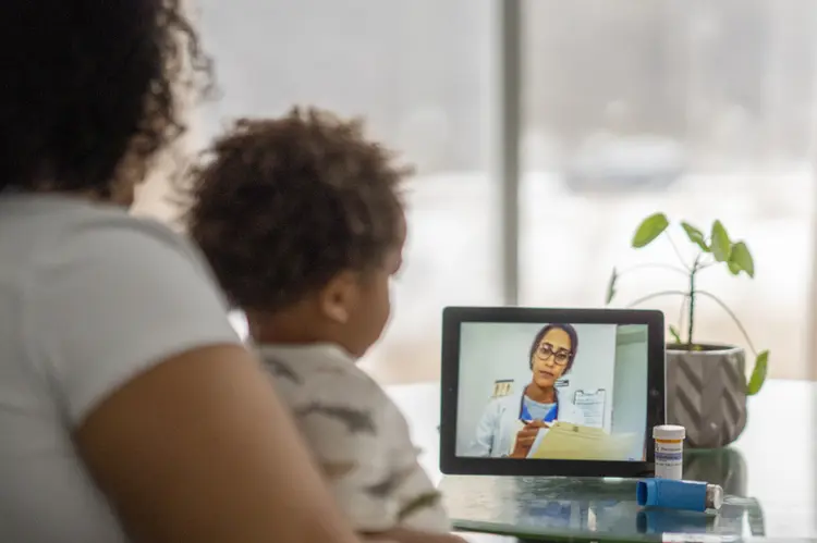 African American mother sitting with her sick child and speaking with a medical professional via video chat during the COVID-19 pandemic. (FatCamera/Divulgação)