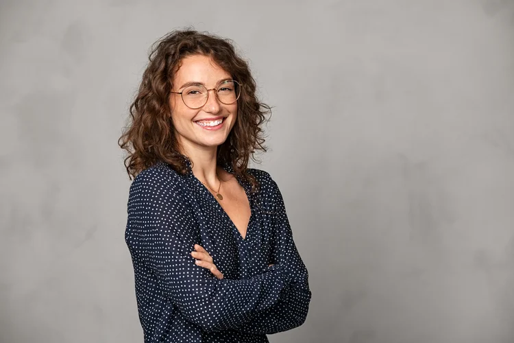 Confident young woman wearing eyeglasses and standing with folded arms on gray wall. Portrait of smiling businesswoman with arms crossed isolated against grey background with copy space. Proud university student girl with specs looking at camera. (Getty Images/Reprodução)