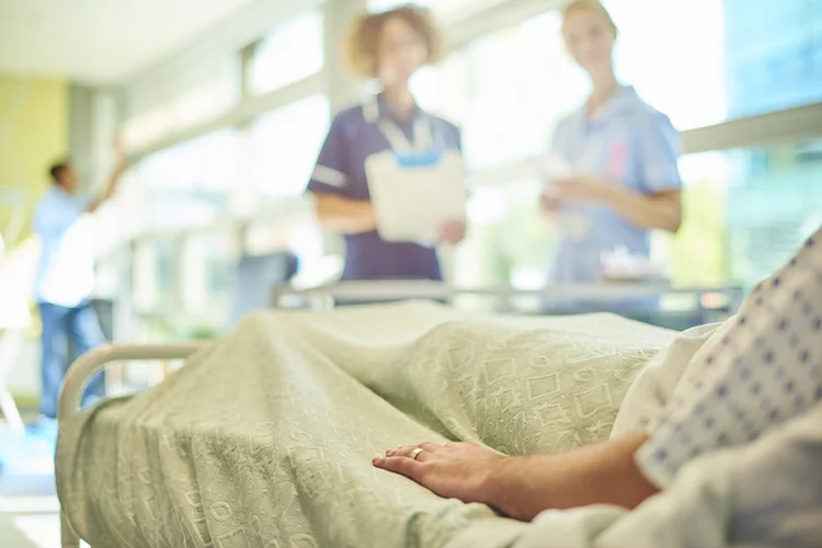 a female senior nurse or matron is  chatting to a young  a young student nurse is attending to the dressings trolley at the foot of a patient's bed . They are all defocussed apart from the patient's hand in the foreground. (sturti/Getty Images)