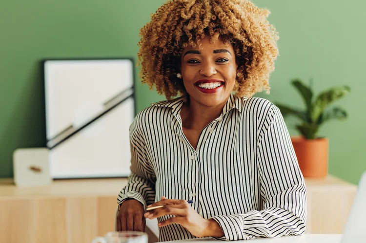 Cheerful African American woman looking at camera while sitting at office desk with laptop computer, mobile phone, notebook and cup of tea. (Getty Images/Getty Images)