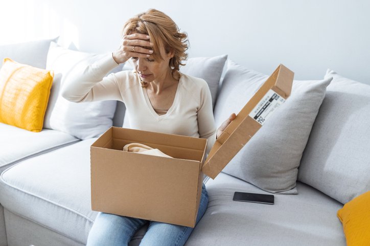 Wrong Item Concept. Sad unpset mature woman sitting on couch and unpacking cardboard box, received parcel with damaged staff, covering face in disappointment, feeling dissatisfied with bad purchase (ljubaphoto/Getty Images)