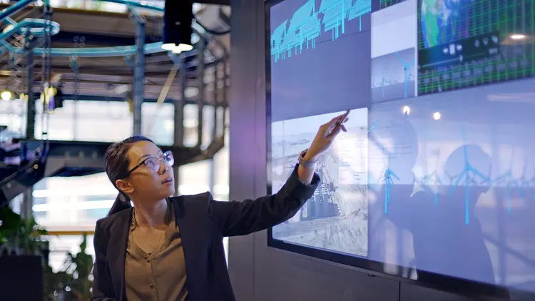 Stock photograph of a young Asian woman conducting a seminar / lecture with the aid of a large screen. The screen is displaying data &amp; designs concerning low carbon electricity production with solar panels &amp; wind turbines. These are juxtaposed with an image of conventional fossil fuel oil production. (iStock/Getty Images)