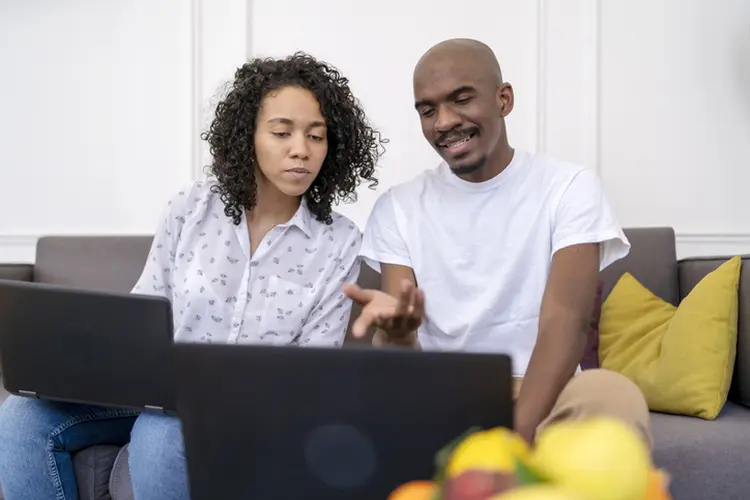 A black couple looking at a laptop screen sitting on the couch in the living room. Happy young African American man and woman discussing something online on a computer. (Getty/Getty Images)