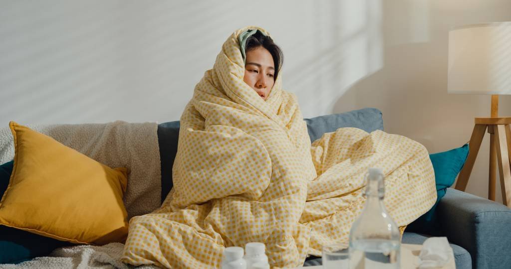 Sick young Asian woman headache fever cough cold sneezing sitting under the blanket on sofa in living room at home. Healthcare concept. (Cris Faga/NurPhoto/Getty Images)