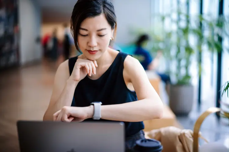 Young Asian businesswoman sitting at her desk in a modern office space, looks down at her watch checking the time while working on laptop. Business and time management concept