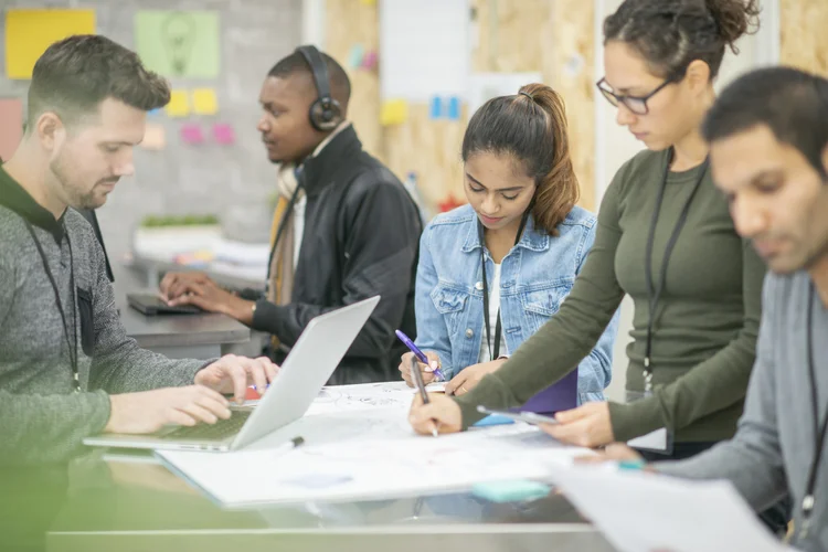 A group of entrepreneurs are indoors in a workshop. They are gathered around a table, and working on blueprints for a new project.