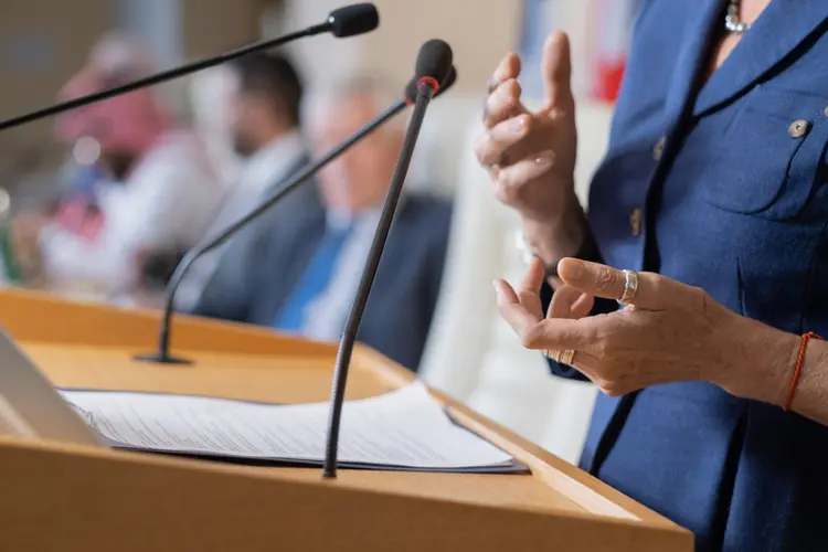Close-up of unrecognizable female politician in rings standing at rostrum with microphones and clipboard while addressing conference (shironosov/Getty Images)