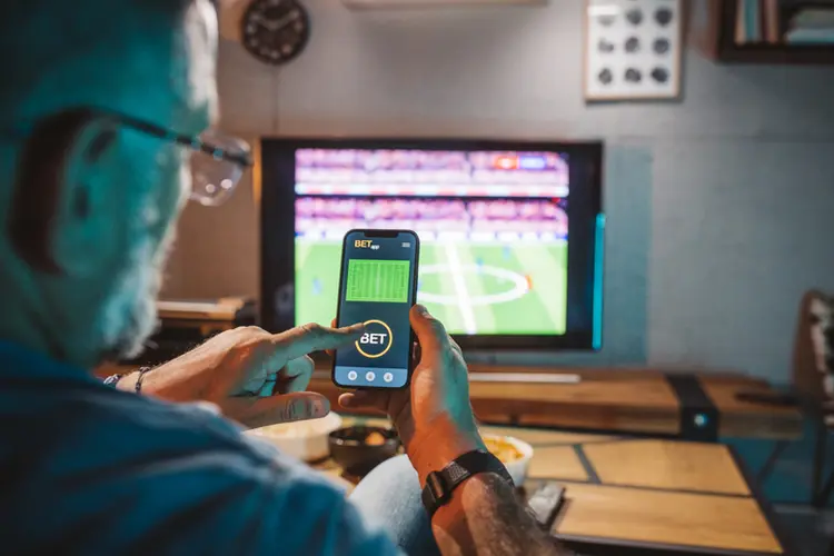 Mature men sitting on sofa at home and watching soccer game.  He is using smart phone for sports betting . (svetkd/Getty Images)