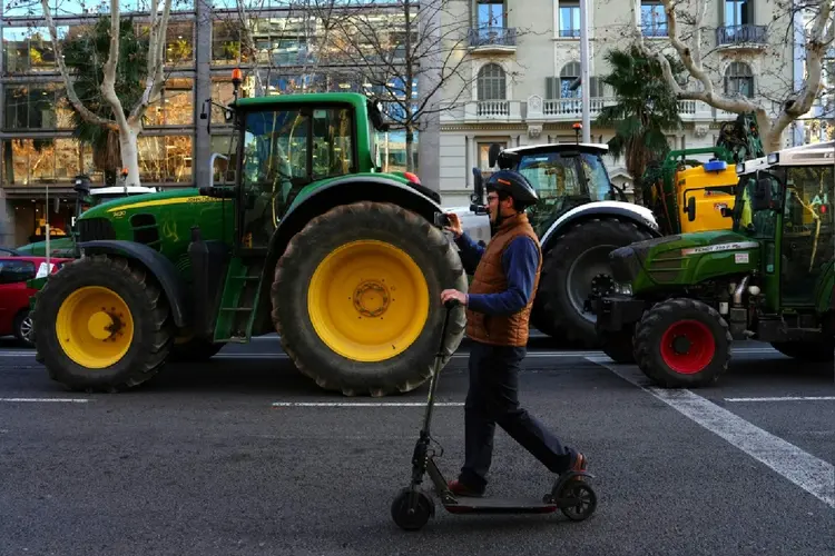 Pessoa passa de patinete em frente a uma fila de tratores concentrados em Barcelona em 7 de fevereiro de 2024 (Valentin Bontemps/AFP Photo)