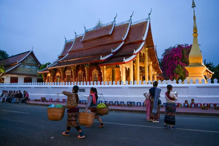 Mulheres carregando cestas ao amanhecer em frente a um templo budista em Luang Prabang, capital turística do Laos. (AFP/Divulgação)