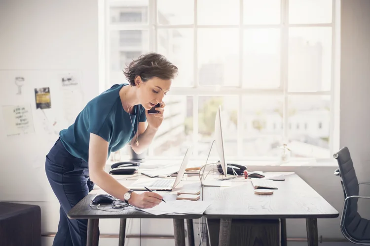 A photo of businesswoman talking on phone while writing on paper at table. Professional is wearing casual. She is in creative office. (Portra/Getty Images)