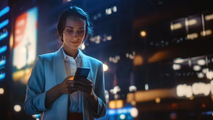 Beautiful Young Woman Using Smartphone Walking Through Night City Street Full of Neon Light. Portrait of Gorgeous Smiling Female Using Mobile Phone. (gorodenkoff/Getty Images)