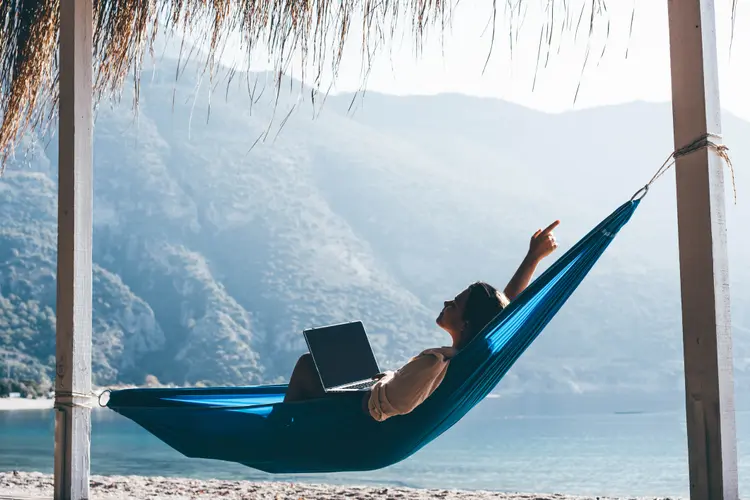 Tourist on summer vacation combining work and leisure. Woman using laptop in hammock on beach. (Maria Korneeva/Getty Images)