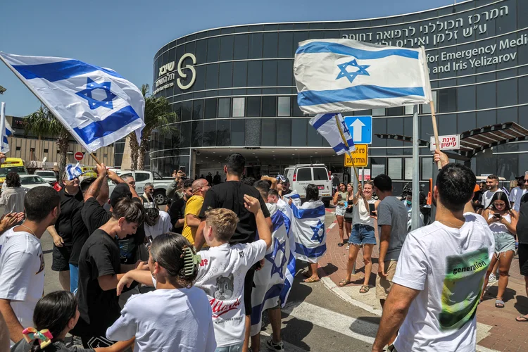 Pessoas se reúnem com bandeiras nacionais israelenses em frente ao Centro Médico Sheba Tel-HaShomer, em Ramat Gan, em 8 de junho de 2024, para onde reféns israelenses foram transferidos após serem resgatados do cativeiro na Faixa de Gaza desde os ataques de 7 de outubro, em meio ao conflito em curso no território palestino entre Israel e o grupo militante Hamas. Israel disse que as suas forças resgataram vivos quatro reféns de um campo de refugiados de Gaza no dia 8 de junho, enquanto intensificavam um ataque, apesar do escrutínio sobre um ataque mortal a uma escola gerida pela ONU naquele local. Os quatro foram sequestrados pelo Hamas no festival de música Nova durante os ataques de 7 de outubro que desencadearam a guerra, disse o exército (JACK GUEZ/AFP)