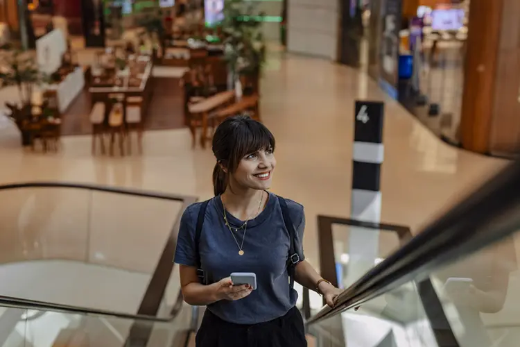 Beautiful young happy Caucasian businesswoman looking away while using a mobile phone and riding an escalator in a mall on her way to work. (miniseries/Getty Images)