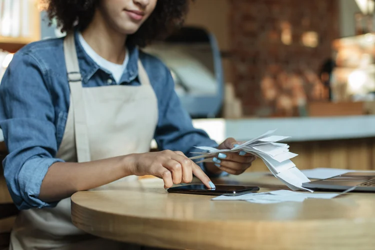 Small business management and accounting. Busy african american young lady owner in apron works with papers, counts costs and profits in interior of modern cafe, cropped, close up, copy space (Prostock-Studio/Getty Images)