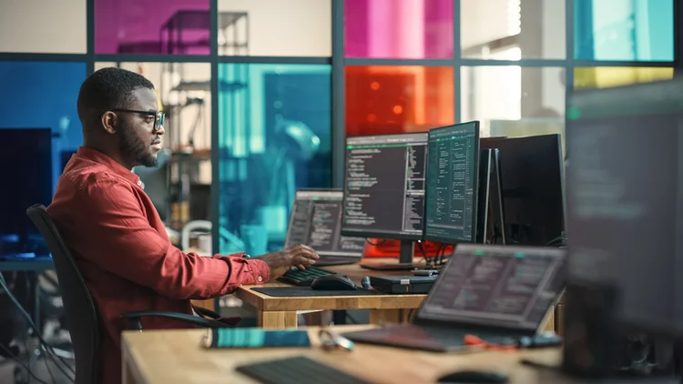 African American Man Writing Lines Of Code On Desktop Computer With Multiple Monitors and Laptop in Creative Office. Male Data Scientist Working on Innovative Online Service For Start-up Company. (gorodenkoff/Getty Images)