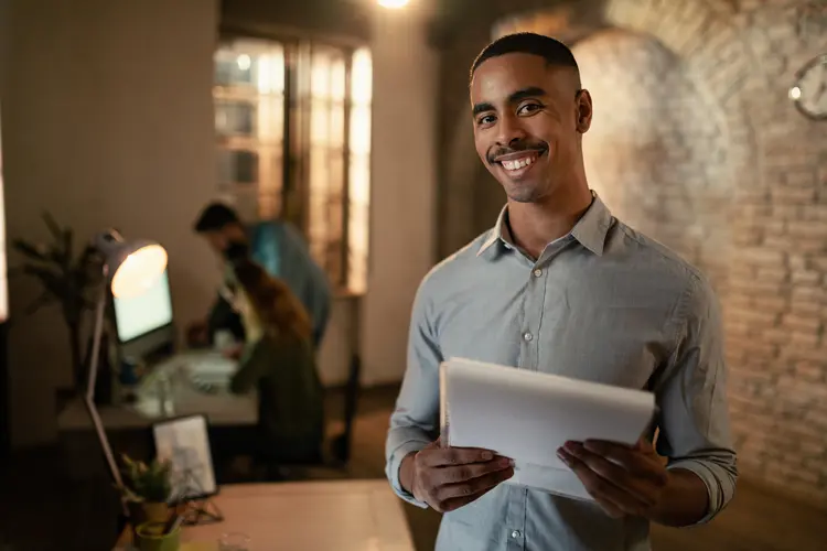 Portrait of happy black entrepreneur working late on business reports and looking at camera in the office.