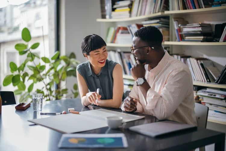 A laughing Japanese businesswoman having a conversation with her African-American coworker while they are sitting at the desk. (FreshSplash/Getty Images)