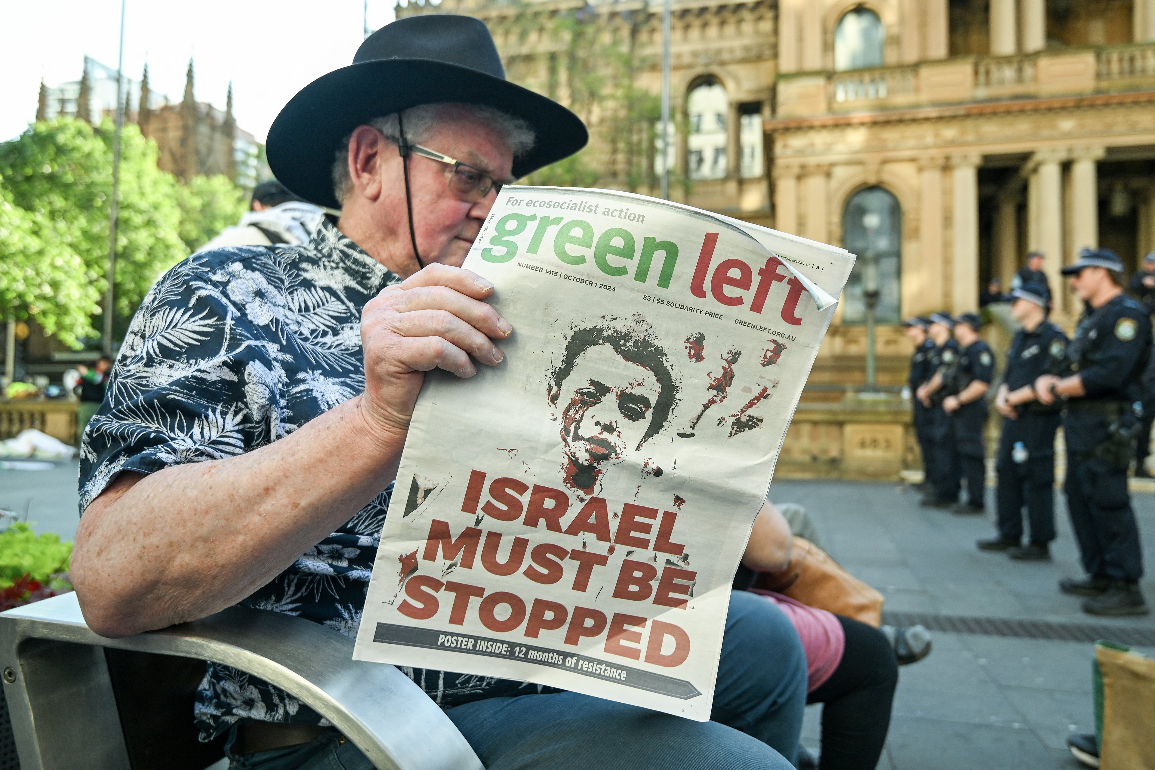 Um homem lê um jornal em um banco enquanto policiais vigiam do lado de fora do Sydney Townhall antes de uma vigília à luz de velas por apoiadores pró-Palestina no primeiro aniversário do conflito Israel-Hamas, em Sydney, em 7 de outubro de 2024