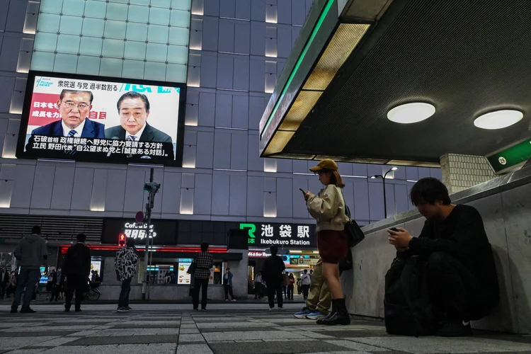 Pessoas ficam do lado de fora da estação de trem de Ikebukuro enquanto imagens do primeiro-ministro japonês Shigeru Ishiba (acima esq.) e do ex-primeiro-ministro e chefe do Partido Democrático Constitucional (CDP) da oposição, Yoshihiko Noda (acima esq.) são transmitidas (Richard A. Brooks /AFP)