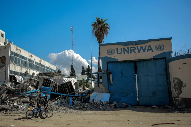 Pessoas passam pela sede danificada da Agência das Nações Unidas de Assistência aos Refugiados da Palestina (UNRWA) na Cidade de Gaza em 15 de fevereiro de 2024, em meio a batalhas em andamento entre Israel e o grupo militante Hamas (AFP)