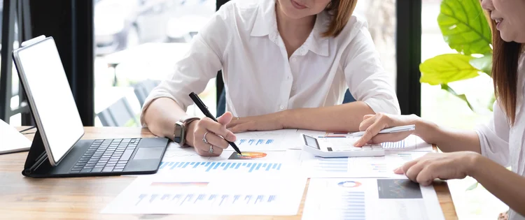 Two women analyzing documents while sitting on a table in office. Woman executives at work in office discussing some paperwork.. (Envato)