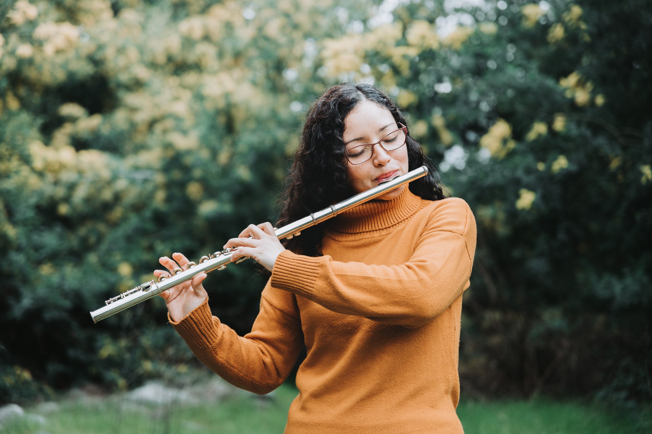 Curly brunette woman with glasses, wearing a mustard yellow sweater, playing transverse flute outdoor in nature