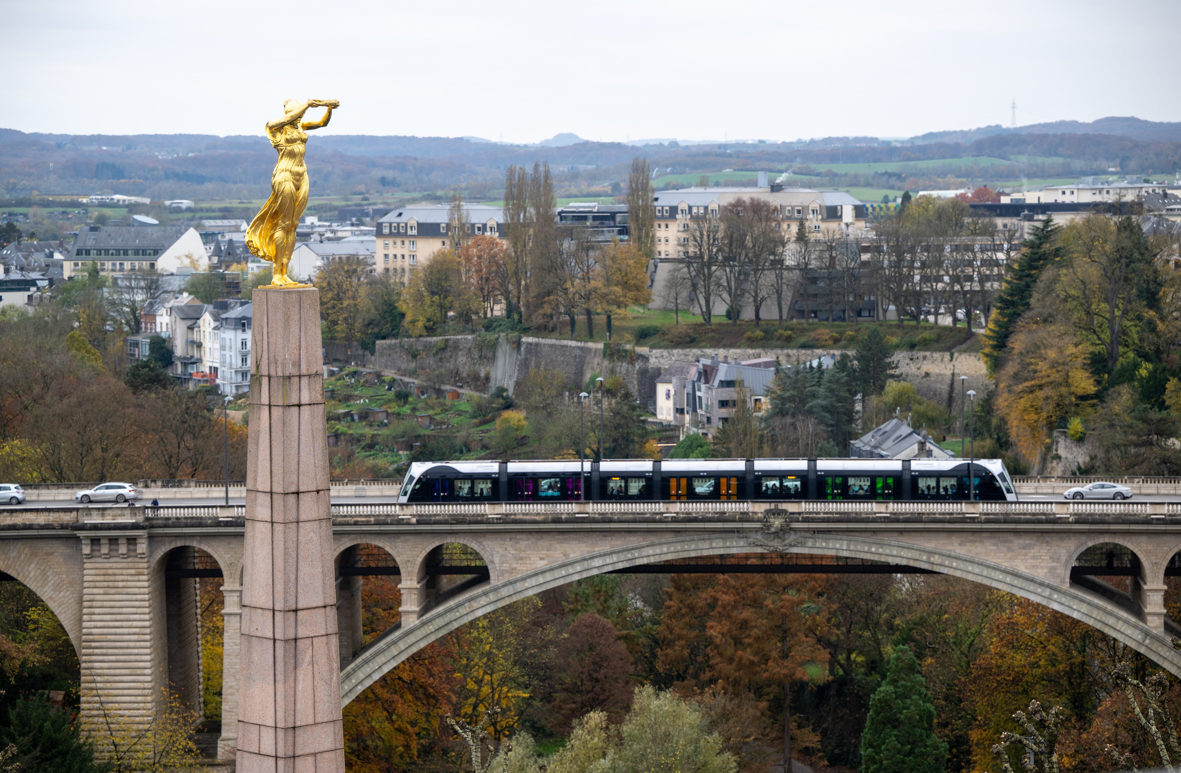 15 November 2024, Luxembourg, Luxemburg: A streetcar crosses the Adolphe Bridge in Luxembourg City. In the foreground is the "Gëlle Fra" (Golden Woman), a so-called war memorial with a gilded bronze statue of a woman on a granite obelisk