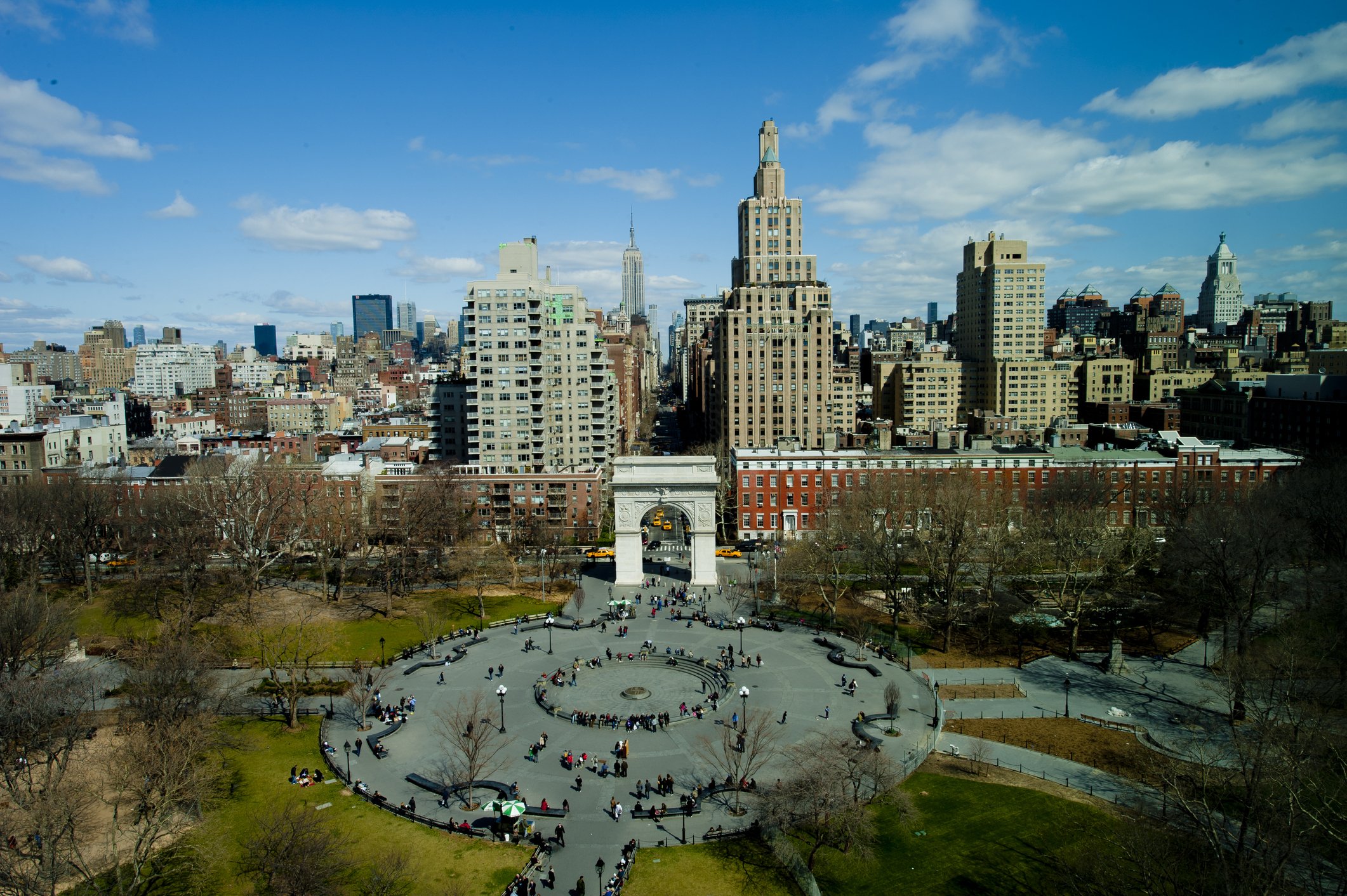 Washington Square Arch at center, while people walk at the square during a sunny day, having the 5th Avenue and Manhattan on the background