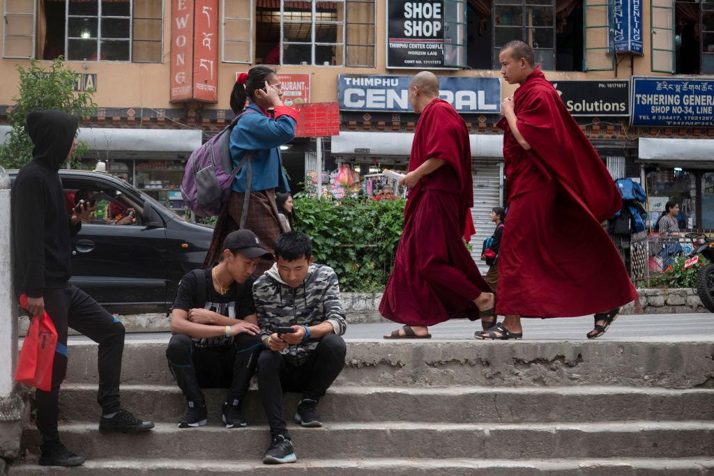 THIMPHU, BHUTAN - JUNE14 : Bhutanese teens are seen hanging out using mobile phones as monks stroll by  on June 14,2018, in Thimphu, Bhutan. While many are expected to wear the 