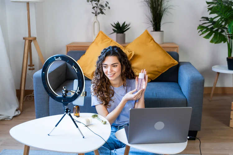 Curly-haired woman showcasing skincare products on camera (PixelVista/Getty Images)