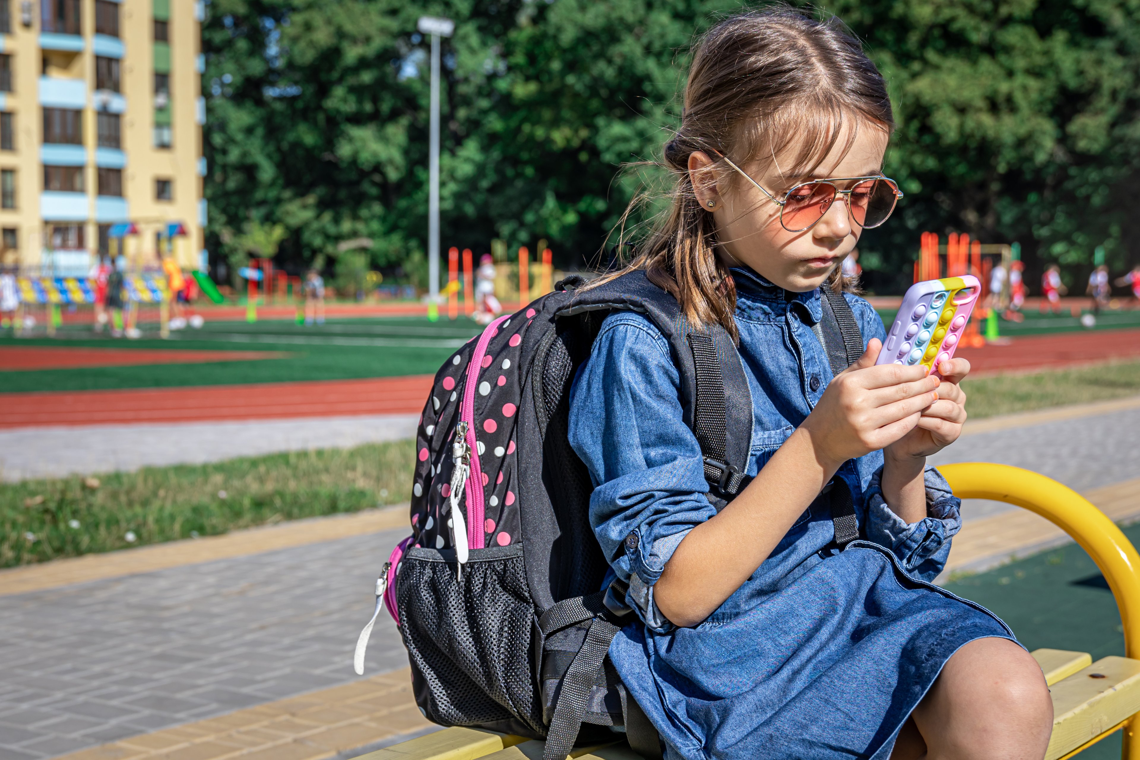 A primary school student with a backpack, uses a smartphone, sitting near the school.