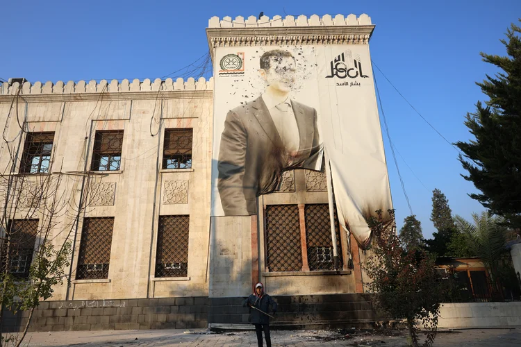 Um combatente anti-regime fica em frente a um retrato danificado do presidente da Síria, Bashar al-Assad, espalhado em um prédio do governo em Hama, um dia depois que os rebeldes capturaram a cidade do centro-oeste da Síria, em 6 de dezembro de 2024 (Mohammed AL-RIFAI/AFP)